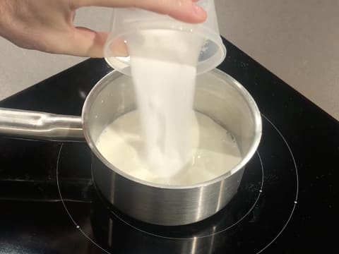 Ajout du sucre en poudre dans le lait qui se trouve dans la casserole posée sur la plaque de cuisson