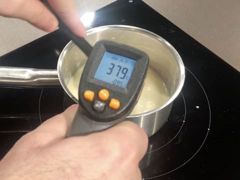 Prise de la température de la préparation lactée qui est mélangée à la spatule maryse dans la casserole, à l'aide d'un thermomètre à visée laser qui affiche 37,9°C