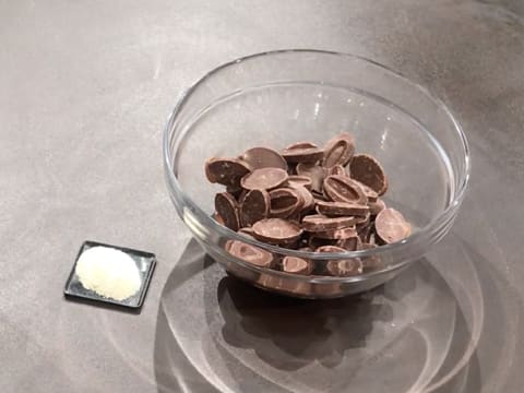 Un saladier en verre posé sur le plan de travail contient des fèves de chocolat au lait et une petite quantité de beurre de cacao Mycryo en poudre est posée à ses côtés, sur le plan de travail