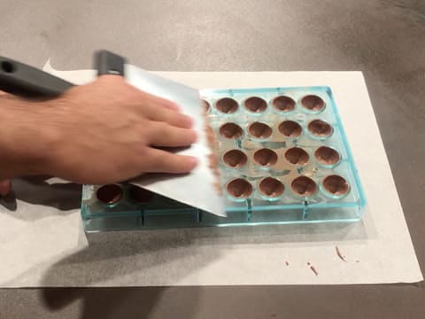 La surface du moule chocolat demi sphère est raclée à l'aide de la spatule à chocolat, le tout posé sur le plan de travail recouvert d'une feuille de papier sulfurisé
