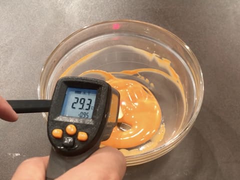 Prise de la température du chocolat blanc fondu coloré en orange dans le saladier en verre à l'aide d'un thermomètre à visée laser qui affiche 29,3°C