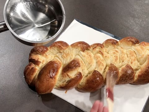 À l'aide d'un pinceau pâtissier, la surface du pain brioché tressé est nappée de sirop de lustrage qui se trouve dans une petite casserole