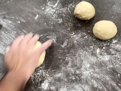 Deux boules de pâte sont sur le plan de travail fariné, et une autre boule de pâte est en train d'être formée à la main