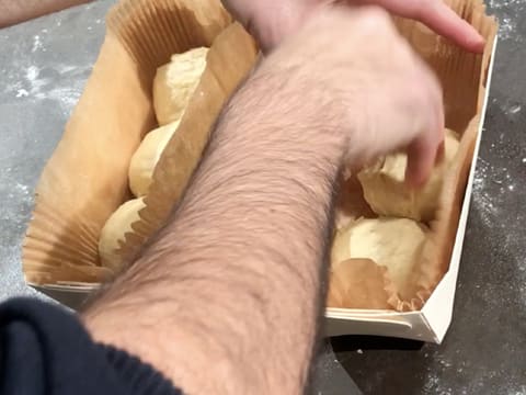 Une barquette de cuisson en bois contient trois boules de pâte à brioche, et une seconde barquette est en train d'être remplie avec deux boules de pâte