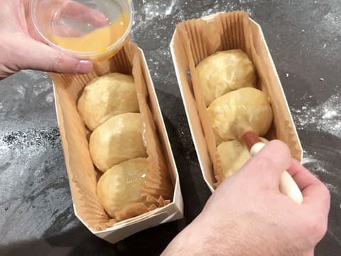 Les boules de pâte à brioche contenues dans les deux barquettes de cuisson en bois, sont dorées avec une dorure et un pinceau pâtissier