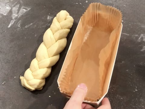 Une barquette de cuisson en bois dans laquelle est déposée une caissette plissée en papier siliconé, est prise dans la main, et la brioche tressée est placée sur le plan de travail