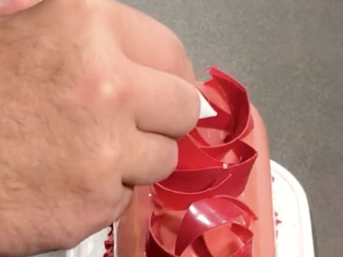 À l'aide d'un cornet décor, des gouttes de nappage miroir neutre sont déposées sur les décors en chocolat à la framboises qui sont sur la bûche glacée