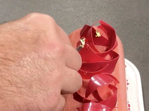 Des pointes de feuille d'or sont déposées sur les gouttes de nappage miroir neutre qui sont sur les décors en chocolat à la framboise, à l'aide d'une pince à dresser