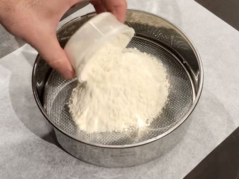La farine est versée dans un tamis qui est posé sur une feuille de papier sulfurisé