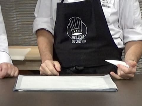 Une plaque à pâtisserie recouverte d'une feuille de papier sulfurisé est placée sur le plan de travail et le Chef tient dans sa main un petit cornet à décor