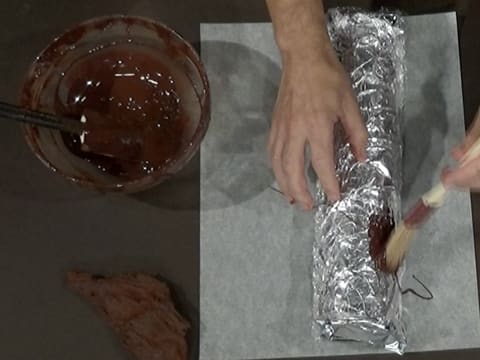 Du chocolat noir fondu est étalé au pinceau pâtissier sur le papier film qui est sur le papier aluminium sur le moule à bûche