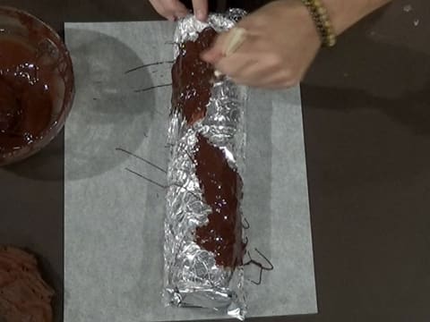 Du chocolat noir fondu est étalé au pinceau pâtissier sur le papier film qui est sur le papier aluminium sur le moule à bûche