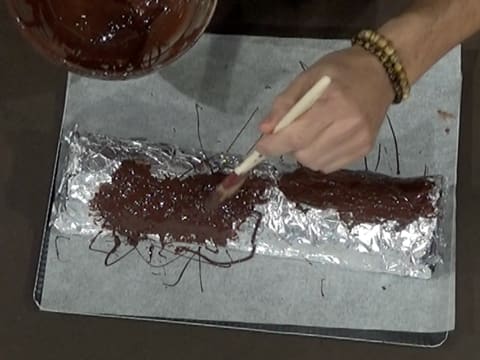 Des fils de chocolat noir fondu sont formés à l'aide du pinceau pâtissier sur le chocolat déjà étalé sur le papier film qui est sur le papier aluminium sur le moule à bûche