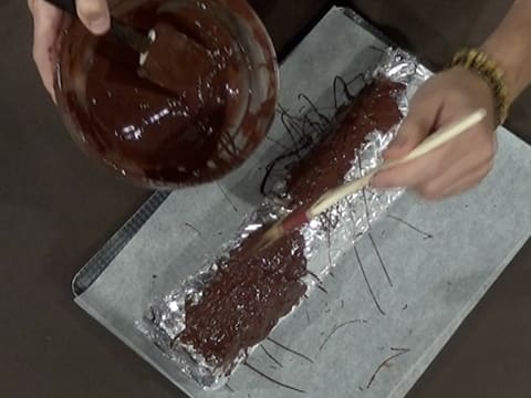 Des fils de chocolat noir fondu sont formés à l'aide du pinceau pâtissier sur le chocolat déjà étalé sur le papier film qui est sur le papier aluminium sur le moule à bûche