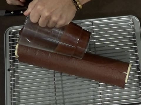 Le glaçage chocolat est versé sur la bûche qui est posée sur la grille à pâtisserie posée sur la plaque creuse