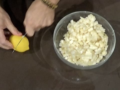 Un citron est coupé en deux au couteau sur le plan de travail, à côté d'un saladier en verre rempli des cubes de poire