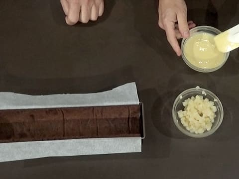 Vue de dessus du moule à bûche chemisé contenant la bande de biscuit chocolat, à côté d'un bol contenant le crémeux poire et d'un autre bol contenant les cubes de poires pochées