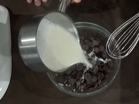 Le lait chaud contenu dans la casserole est versé sur les pistoles de chocolat noir contenues dans un saladier en verre