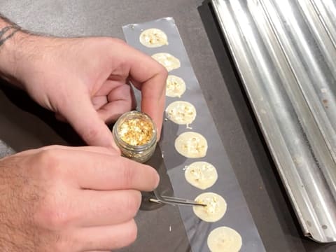 Des paillettes d'or sont déposées sur les pastilles de chocolat blanc, à l'aide d'une pince à dresser