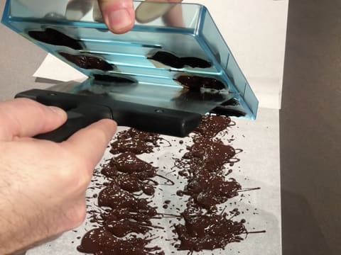 Le moule est arasé avec la spatule à chocolat et le chocolat noir tempéré coule sur le plan de travail qui est recouvert d'une feuille de papier sulfurisé