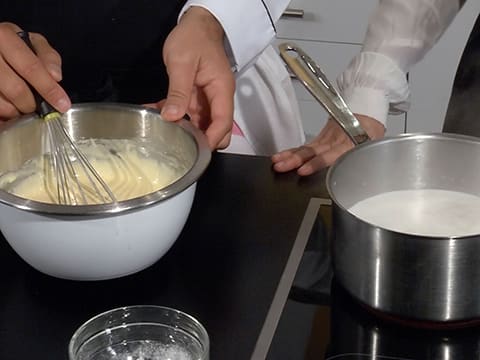 La préparation dans le cul de poule est mélangée au fouet et à côté le lait est en train de cuire dans la casserole qui est placée sur la plaque de cuisson