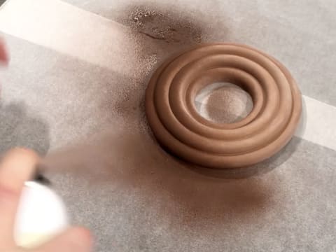 Du colorant en spray effet velours brun est pulvérisé sur l'anneau de crémeux au chocolat qui est posé sur le plan de travail recouvert de feuilles de papier sulfurisé