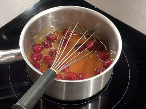 La purée de mangue et les griottes sont en train de cuire dans la casserole qui contient aussi un fouet et qui est placée sur une plaque de cuisson