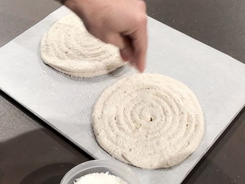 De la noix de coco râpée est parsemée sur deux disques de dacquoise pochés sur une feuille de papier sulfurisé posée sur une plaque à pâtisserie