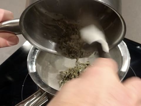 La crème infusée au jasmin dans la casserole est filtrée dans une passoire fine au-dessus de la casserole contenant le lait sur la plaque de cuisson