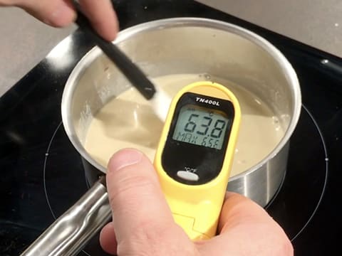 Prise de la température de la préparation crémeuse dans la casserole qui est sur la plaque de cuisson, à l'aide d'un thermomètre à visée laser qui affiche 63,8°C