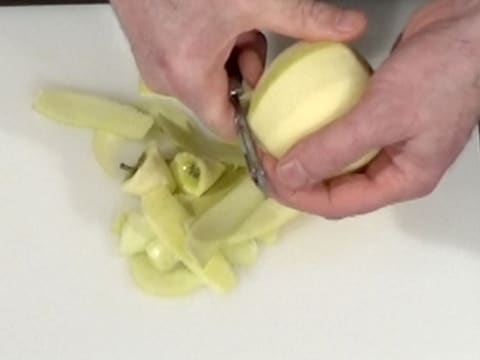 Une pomme est pelée au dessus d'une planche à découper, à l'aide d'un éplucheur