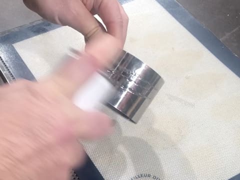 Un cercle à mousse est graissé à l'aide d'un spray de démoulage, au dessus d'un tapis de cuisson en silicone