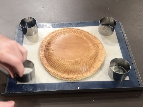 Quatre petits cercles à mousse sont placés au quatre coins de la plaque à pâtisserie qui est recouverte du tapis en silicone sur lequel se trouve la galette des rois décorée