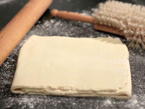 Obtention de la pâte feuilletée inversée sur le plan de travail fleuré, à côté du rouleau à pâtisserie et de la brosse à farine