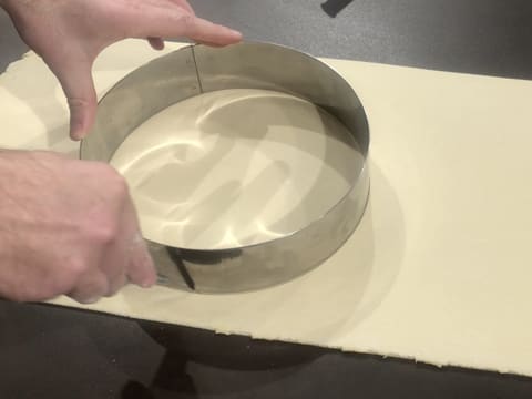 Un cercle à mousse est posé sur l'abaisse de pâte feuilletée inversée et un disque de pâte est détaillé avec un couteau qui suit le contour du cercle inox