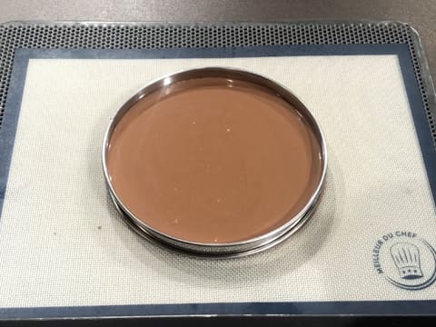 Obtention d'un cercle à tarte inox qui contient du gianduja et qui est placé sur une plaque de cuisson perforée recouverte d'un tapis silicone, le tout posé sur le plan de travail