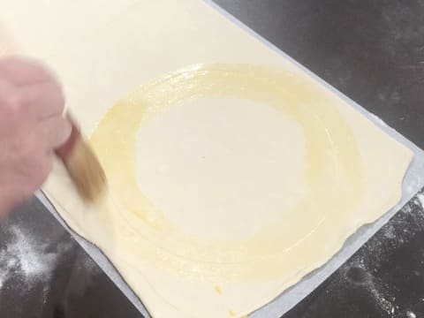 De la dorure est étalée avec un pinceau à pâtisserie sur le tracé du disque sur l'abaisse de pâte feuilletée