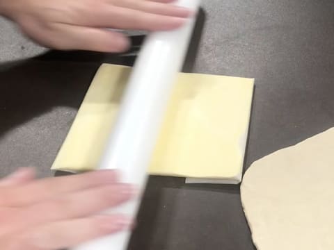 Le beurre de tourage qui est enveloppé dans une feuille de papier sulfurisé est étalé sur le plan de travail à l'aide du rouleau à pâtisserie