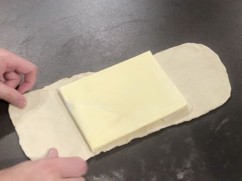 Le beurre de tourage qui est enveloppé dans une feuille de papier sulfurisé et qui est abaissé en un rectangle, est posé au milieu de la pâte étalée en une bande sur le plan de travail
