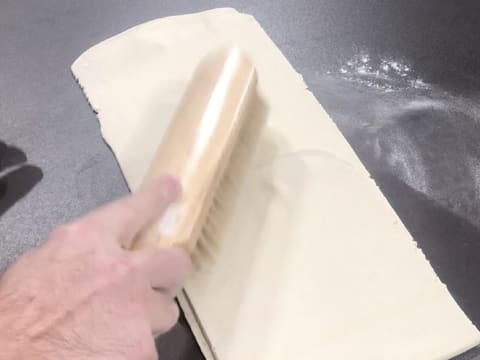 L'excédent de farine est retiré avec une brosse à farine qui est passée sur le rectangle de pâte posé sur le plan de travail fleuré