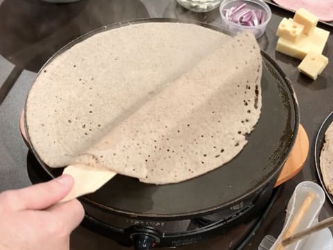 La galette qui est en train de cuire sur la crêpière électrique est soulevée avec une spatule à crêpes en bois