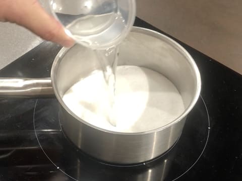 L'eau est versée sur le sucre en poudre qui est dans une casserole posée sur la plaque de cuisson