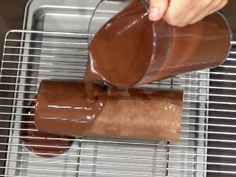 Le gâteau roulé qui est sur la grille et sur une plaque creuse, est nappé de glaçage chocolat