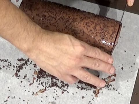 La bûche au chocolat qui est posée sur une feuille de papier sulfurisé, a une de ses extrémités qui est parée avec un couteau