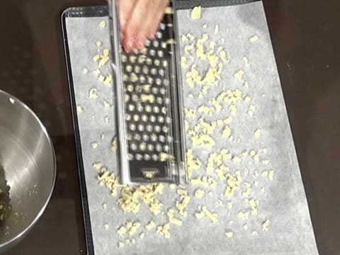 La pâte à crumble est râpée sur une grille à spätzle et des copeaux de pâte tombent sur une feuille de papier sulfurisé qui est placée sur une plaque à pâtisserie sur le plan de travail
