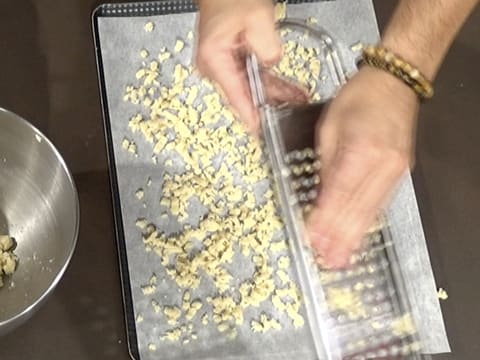 La pâte à crumble est râpée sur une grille à spätzle et des copeaux de pâte tombent sur une feuille de papier sulfurisé qui est placée sur une plaque à pâtisserie sur le plan de travail