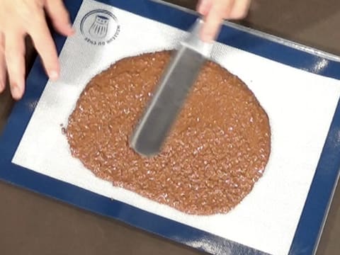 La pâte à croustillant chocolat est étalée sur le tapis de cuisson en silicone, à l'aide d'une spatule métallique coudée