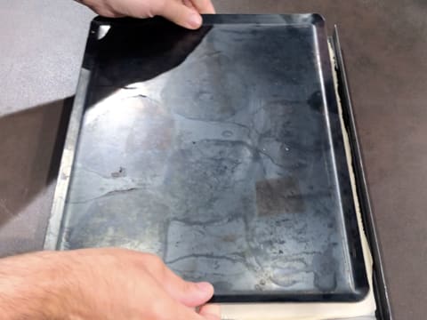 L'abaisse de pâte feuilletée recouverte d'une feuille de papier sulfurisé est recouverte d'une plaque à pâtisserie en tôle bleuie