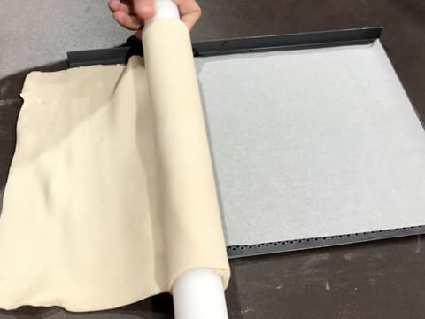 La pâte feuilletée enroulée autour du rouleau à pâtisserie, est déroulée sur une plaque à pâtisserie perforée recouverte d'une feuille de papier sulfurisé