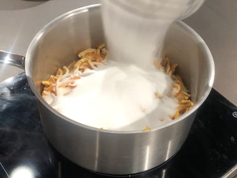 Ajout du sucre en poudre sur les lanières d'écorce et les segments des clémentines, dans la casserole qui est posée sur la plaque de cuisson